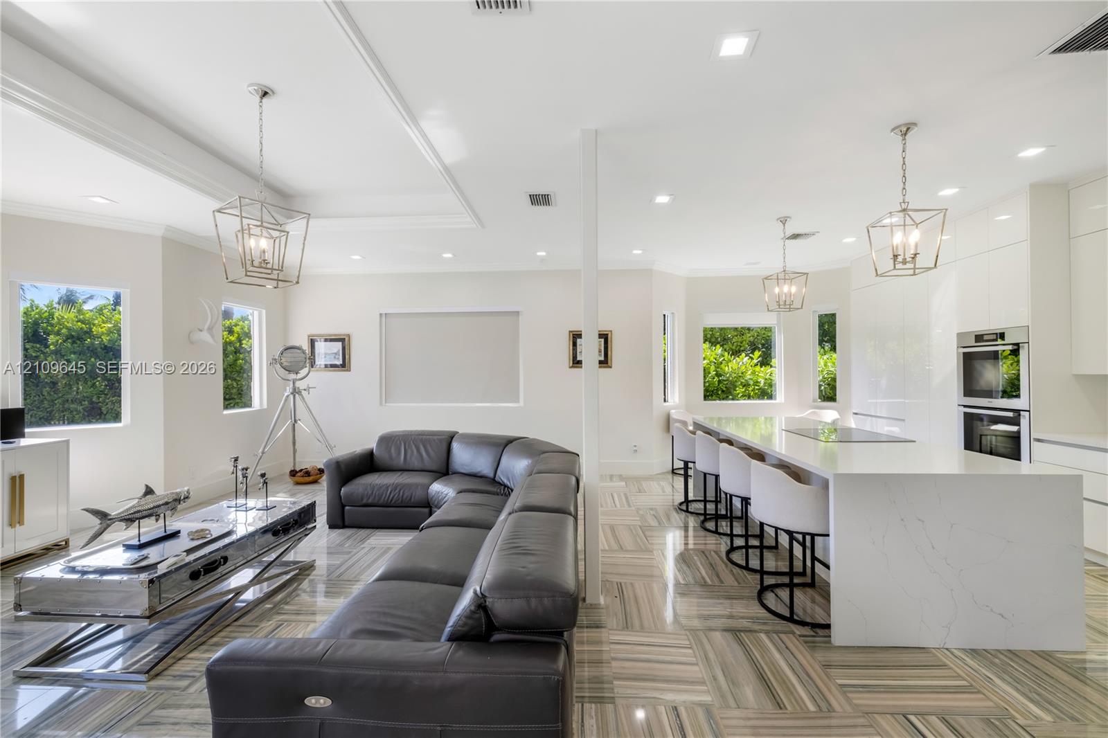 Open-concept interior featuring large-format tile flooring, a waterfall-edge stone island with integrated cooktop, dual built-in wall ovens, recessed lighting, and geometric metal pendant fixtures