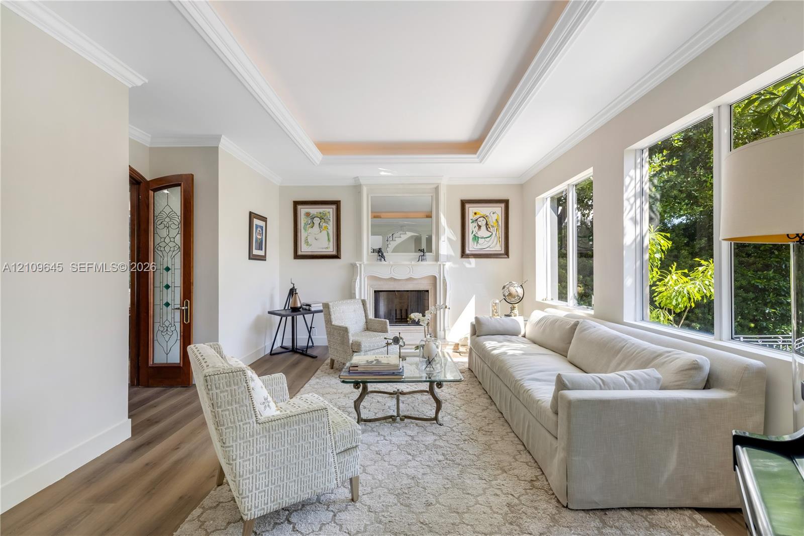 Living area featuring wood-finish flooring, a tray ceiling with crown molding, a wood-burning fireplace with a mantel, and oversized windows
