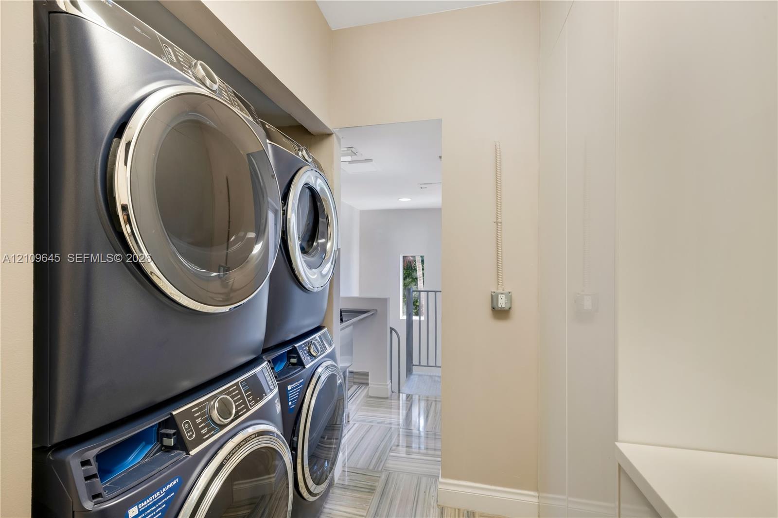 Laundry area featuring stacked metallic-finish washing machine and dryer, floor-to-ceiling built-in cabinetry, and neutral stone-patterned tile flooring