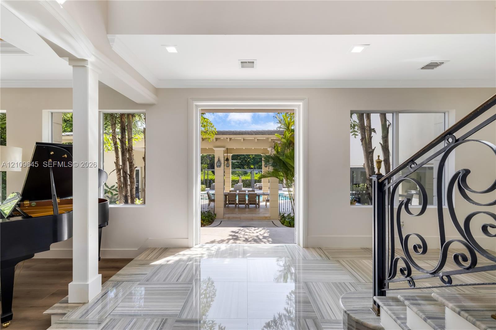 Foyer featuring large-format light-toned stone flooring, decorative iron stair railing, recessed ceiling lighting, and expansive square-cased entry to the outdoor patio