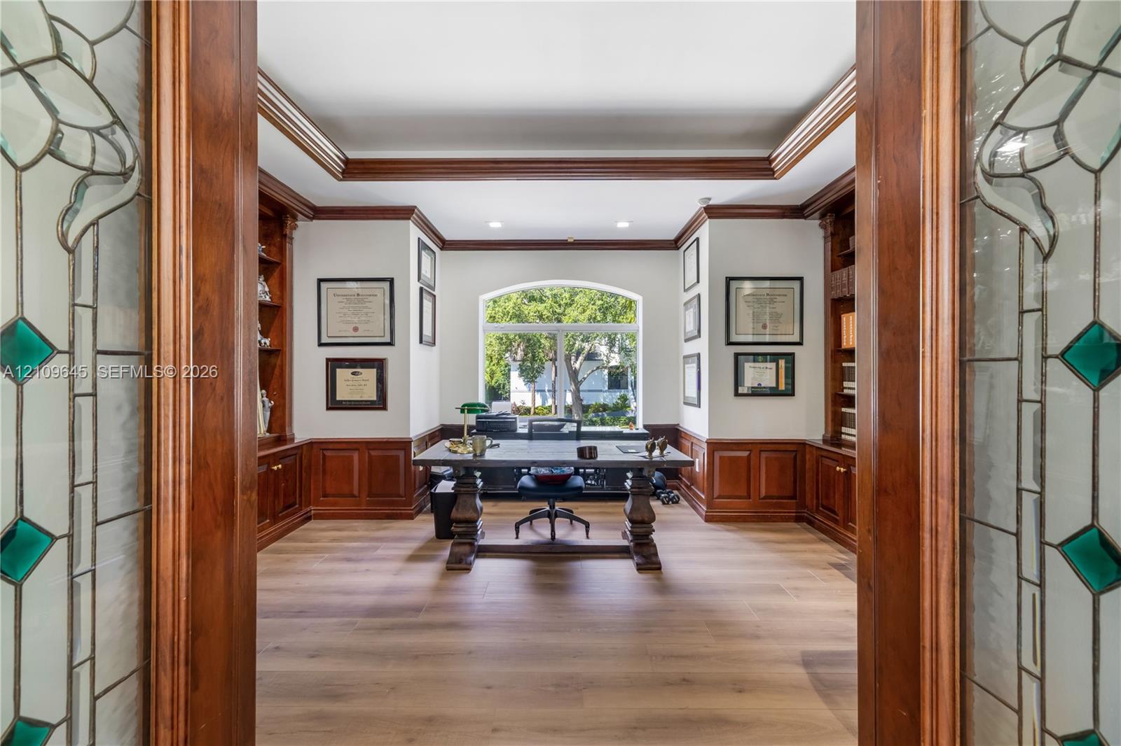 Custom woodwork study featuring wood-paneled wainscoting, floor-to-ceiling built-in cabinetry, arched window, recessed lighting, and glass-paneled double doors with decorative detailing