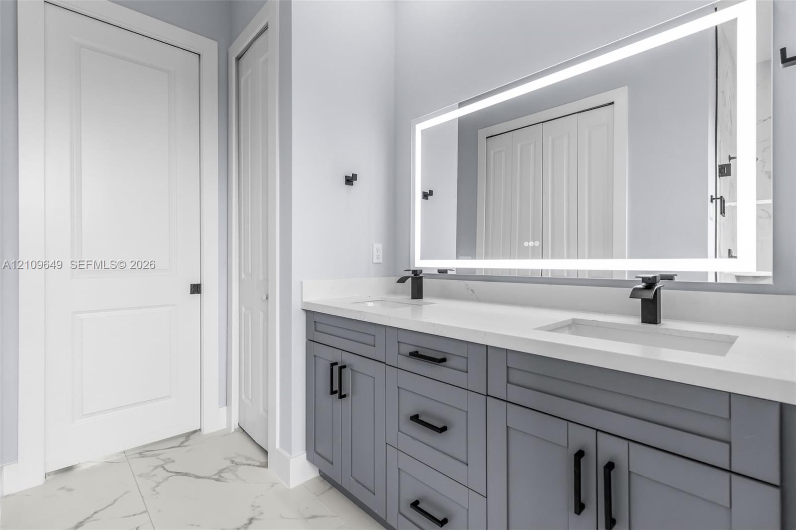 Double vanity with grey shaker-style cabinetry, white solid-surface countertop, integrated dual sinks, matte black fixtures, and a large backlit rectangular mirror