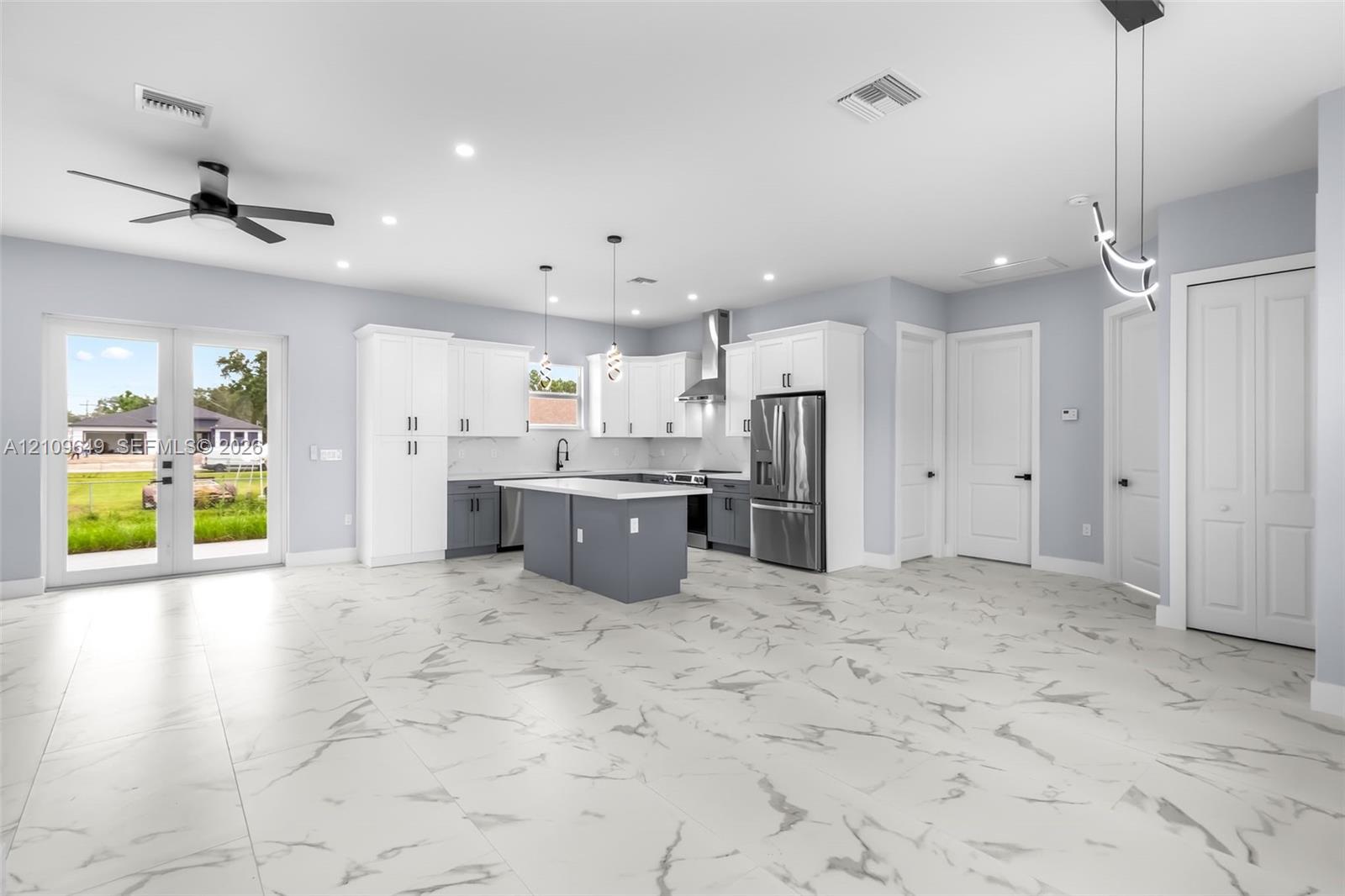 Open-concept kitchen featuring two-toned cabinetry, a central island with a stone-finish countertop, stainless steel appliances, porcelain tile flooring, and glass-paneled double doors