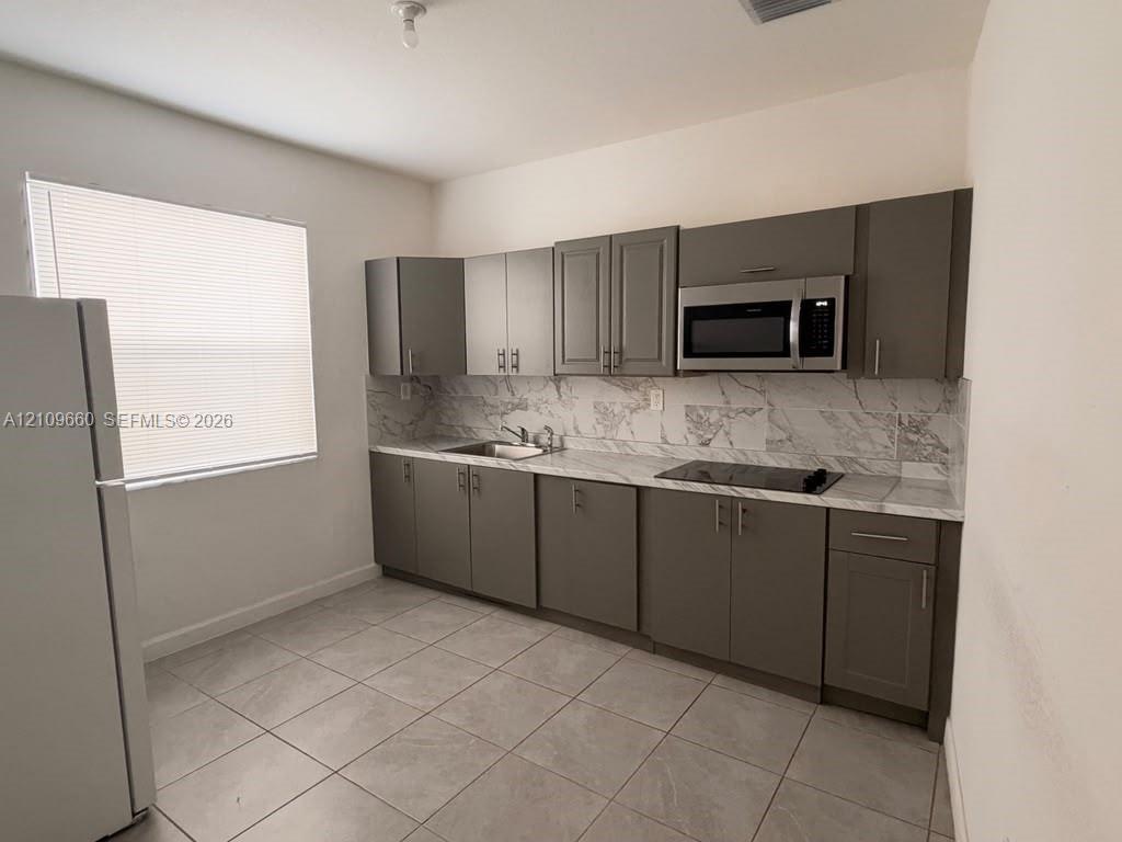 Kitchen featuring gray shaker-style cabinetry, stone-patterned backsplash, light-toned countertop, built-in microwave, and tile flooring Kitchen featuring gray shaker-style cabinetry, stone-patterned backsplash, light-toned countertop, built-in microwave, and tile flooring