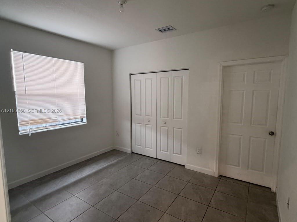 Room featuring neutral-toned floor tiling, a multi-panel door, built-in closet with double doors, and a window with horizontal blinds Room featuring neutral-toned floor tiling, a multi-panel door, built-in closet with double doors, and a window with horizontal blinds