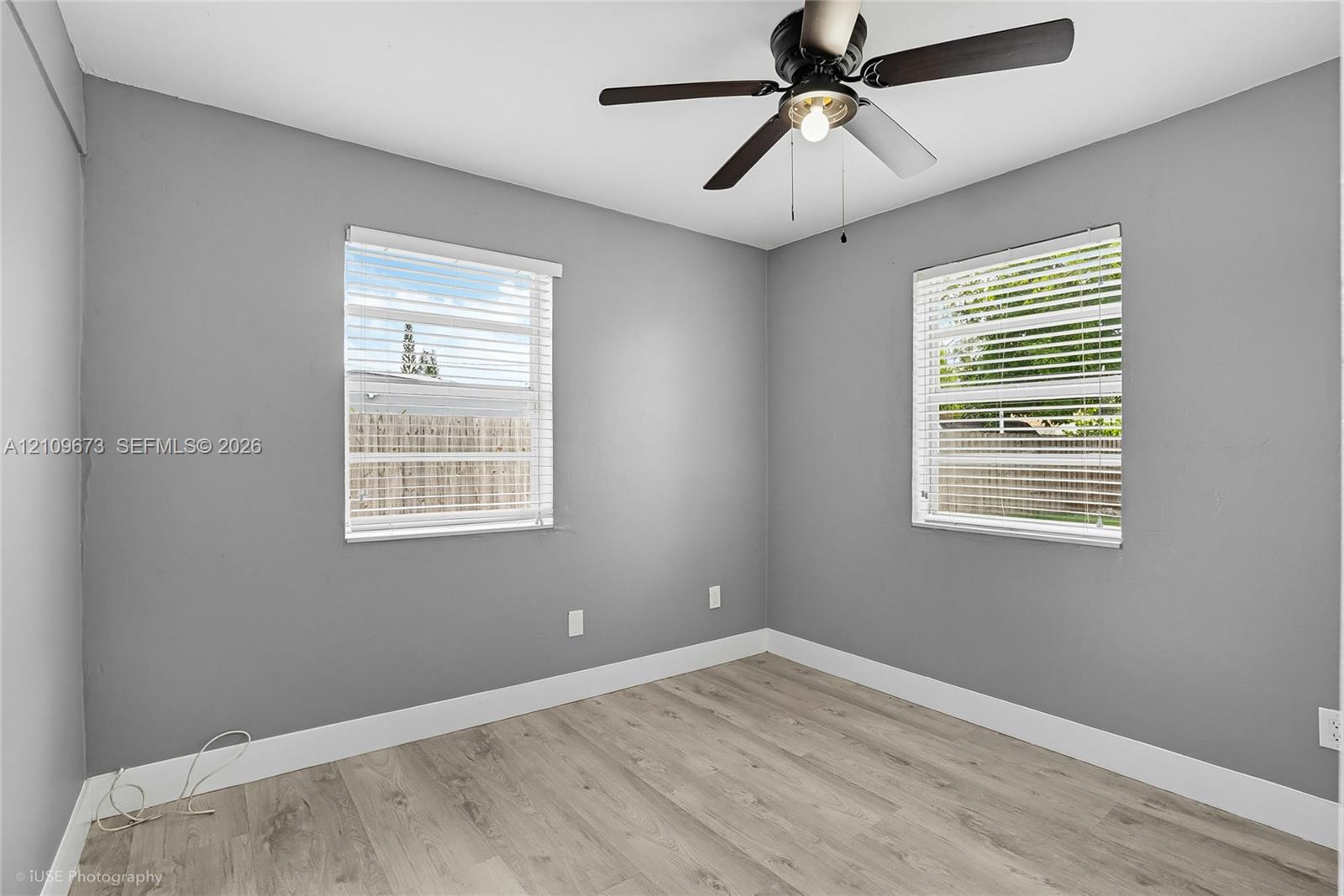 Interior space featuring wood-finish flooring, neutral gray wall paint, dual windows with horizontal blinds, and a central ceiling fan with an integrated light fixture