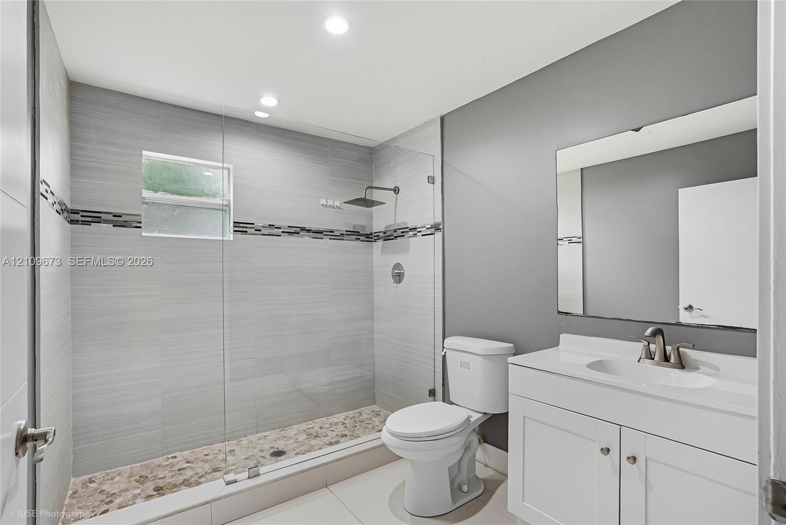 Walk-in shower with floor-to-ceiling gray tile, pebble-stone flooring, frameless glass enclosure, and a rain-style showerhead, complemented by a white vanity with an integrated sink and rectangular wall mirror