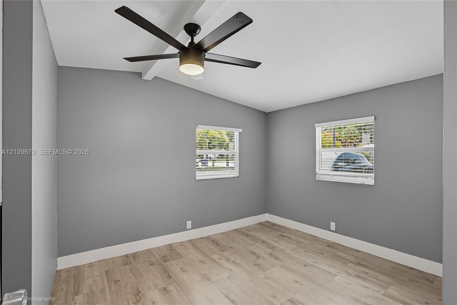 Room featuring wood-finish flooring, vaulted ceilings with an exposed beam, a matte black ceiling fan with integrated lighting, and dual windows with horizontal blinds