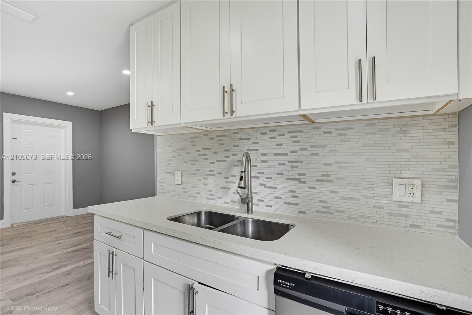 White shaker-style cabinetry, solid surface countertop, stainless steel undermount double-basin sink with gooseneck faucet, horizontal tile backsplash, and wood-finish flooring