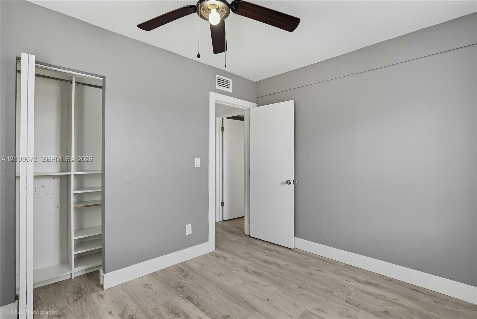 Neutral-toned interior featuring wood-finish flooring, a built-in closet with multi-tier shelving, and a ceiling-mounted fan with integrated lighting
