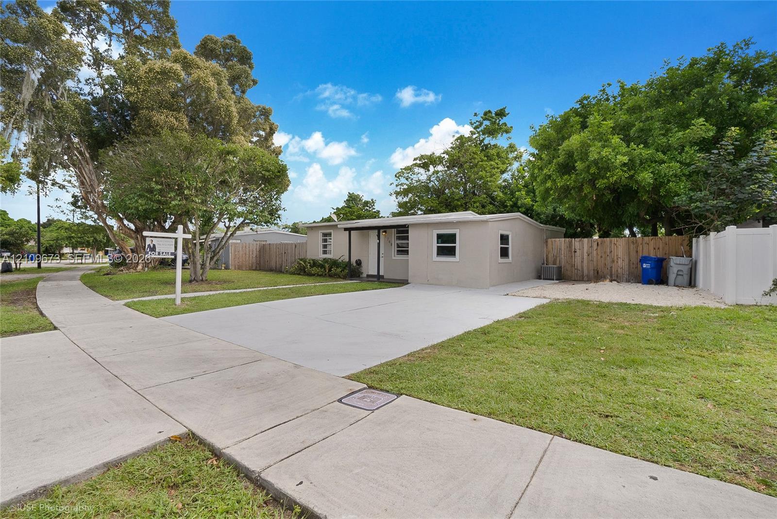 Single-story exterior featuring a concrete driveway, lawn, mature trees, and wood-panel perimeter fencing Single-story exterior featuring a concrete driveway, lawn, mature trees, and wood-panel perimeter fencing