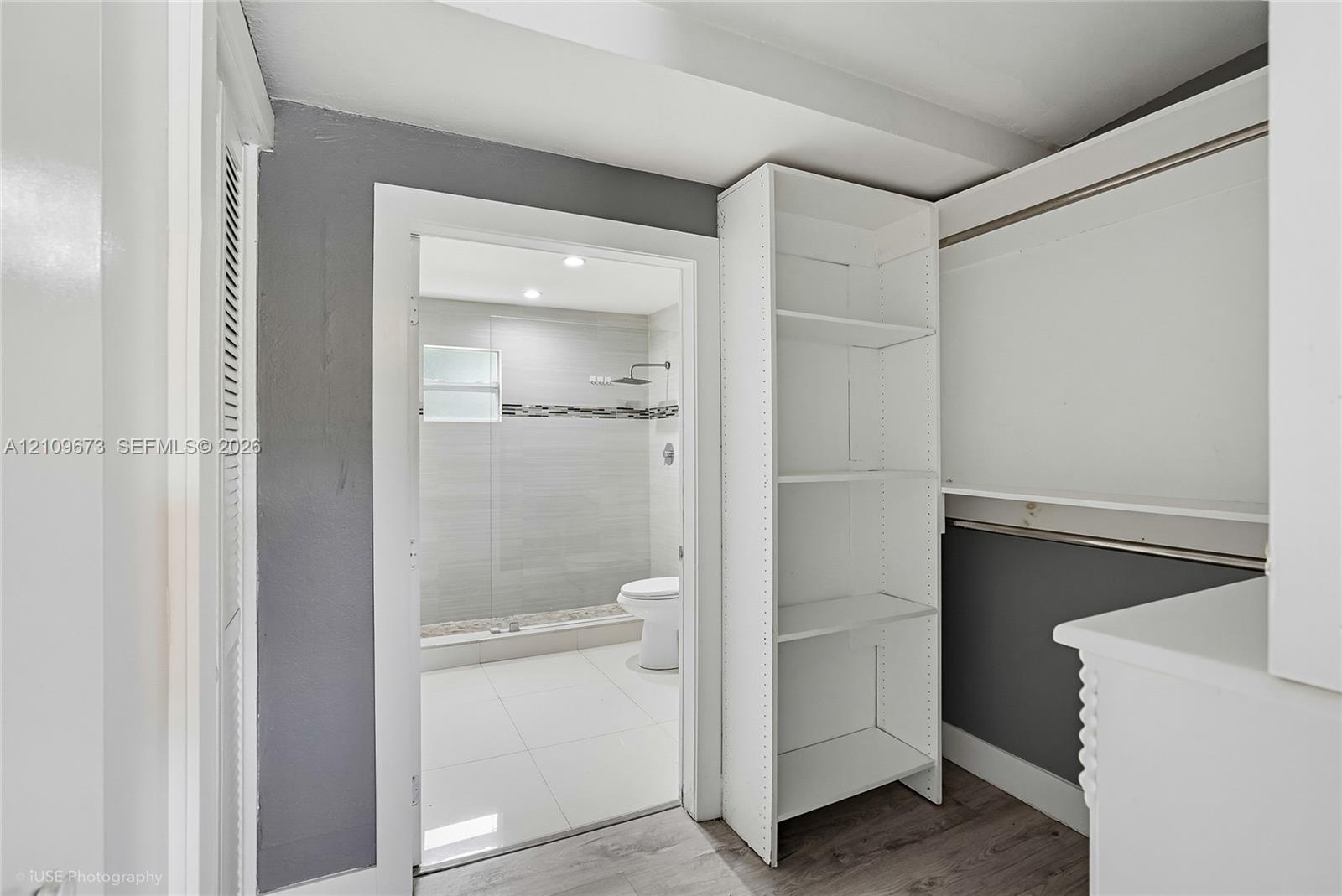 Walk-in storage area featuring custom white built-in shelving, wood-finish flooring, and a doorway revealing a tiled bathroom with a glass-enclosed shower, rain showerhead, and stone-accented wall detailing