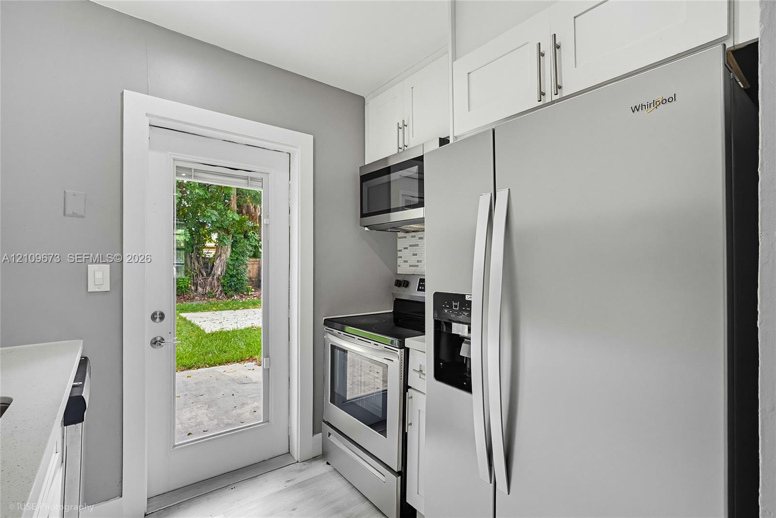 Kitchen area featuring white shaker-style cabinetry, stainless steel appliances, light-toned wood-finish flooring, and a glass-paneled exterior door Kitchen area featuring white shaker-style cabinetry, stainless steel appliances, light-toned wood-finish flooring, and a glass-paneled exterior door