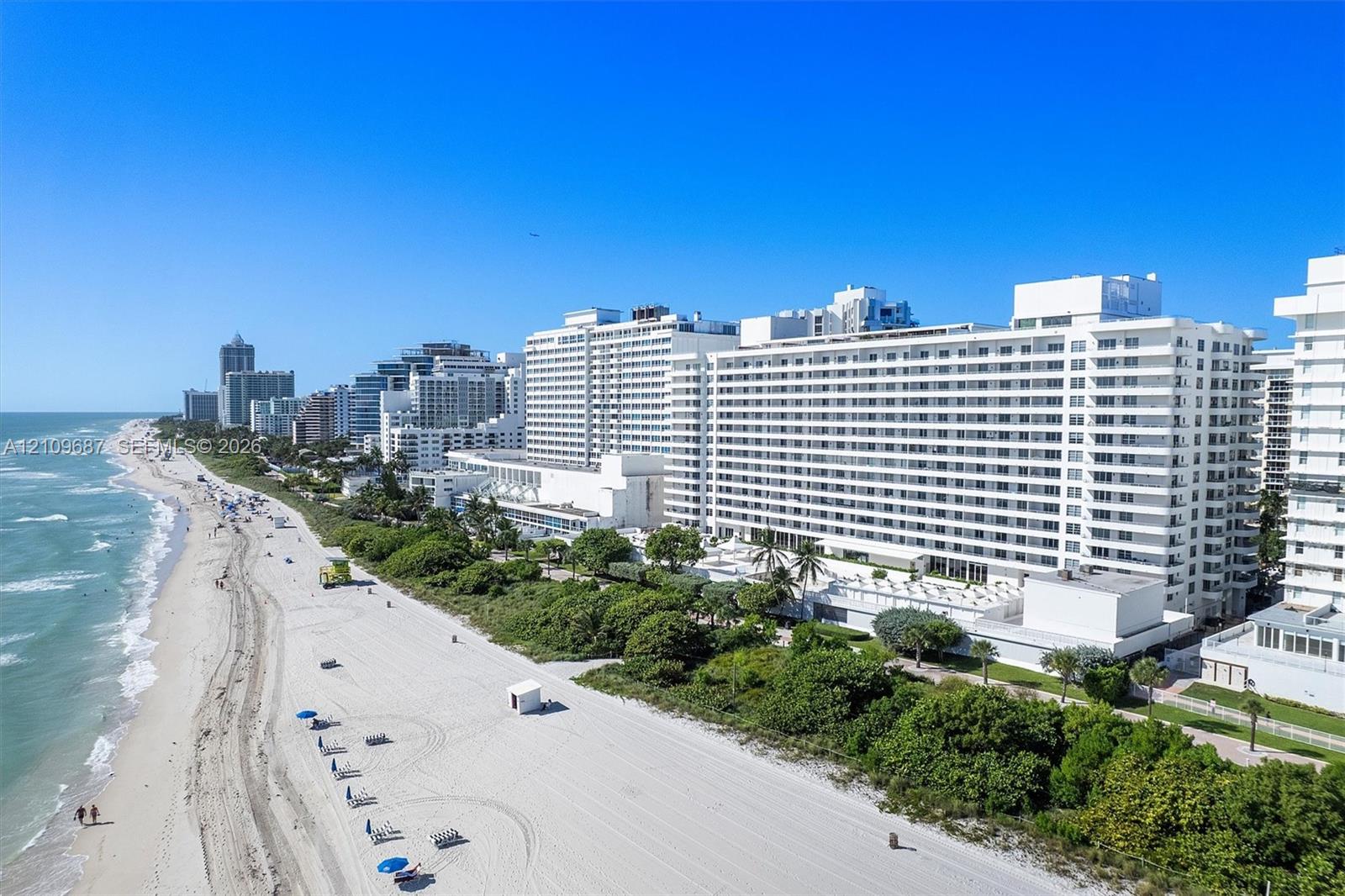 Coastal high-rise building with white stucco facade, expansive balcony tiers, direct beachfront access, and mature landscaping