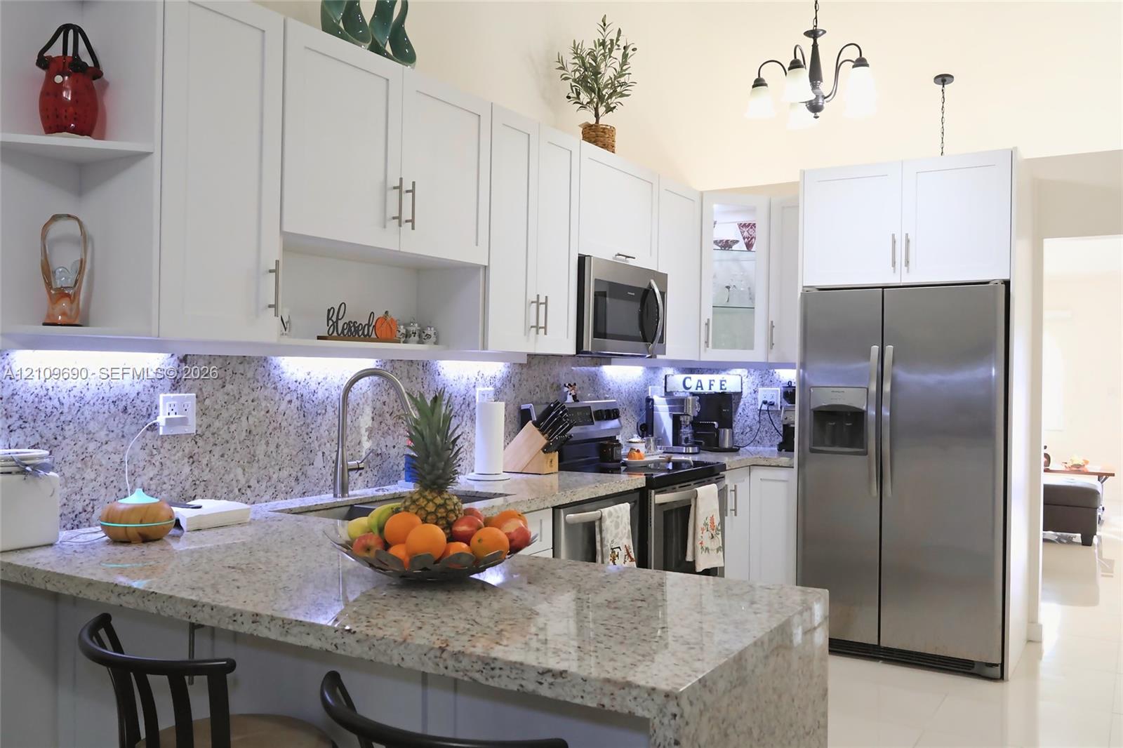 Kitchen featuring white shaker-style cabinetry, stone countertops, stainless steel appliances, under-cabinet lighting, and a multi-arm chandelier