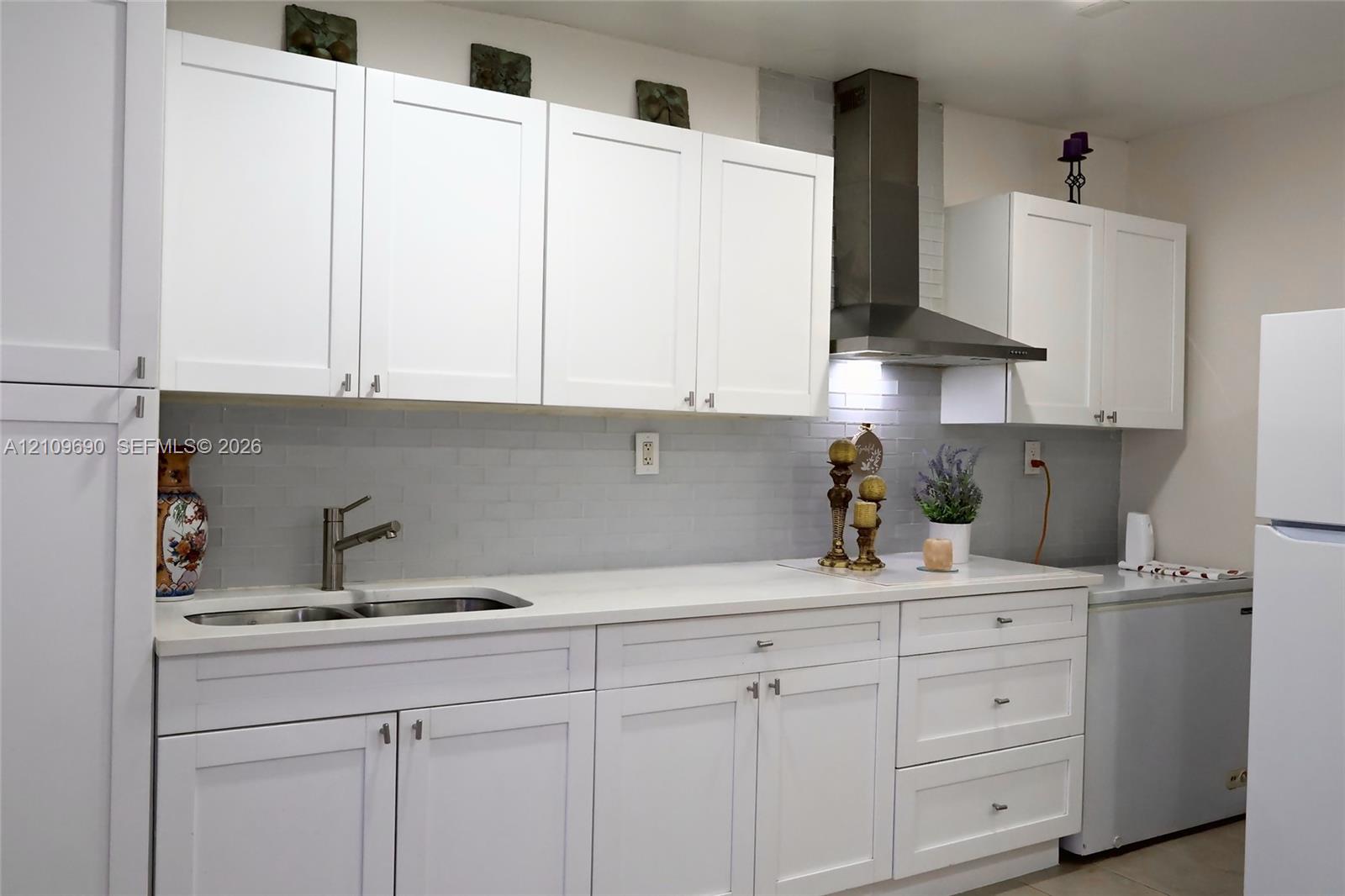 Kitchen featuring white shaker-style cabinetry, a light-toned countertop, a stainless steel chimney-style range hood, and a light gray tile backsplash