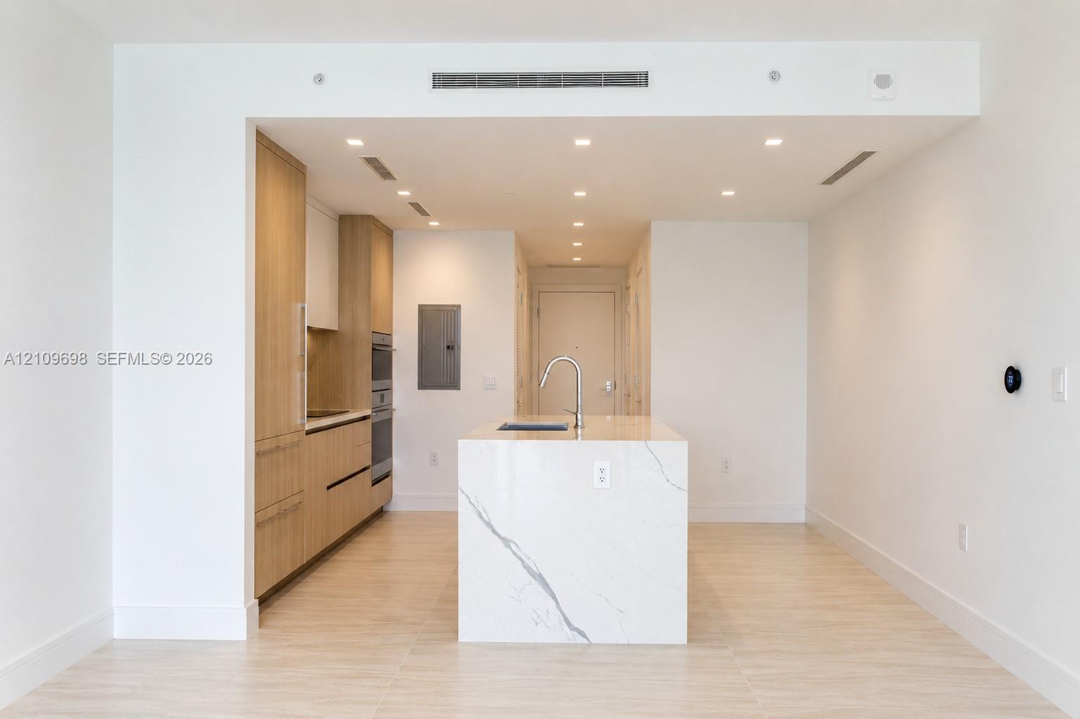 Modern kitchen featuring light wood-finish cabinetry, a central island with a stone-finish countertop, stainless steel wall-mounted oven, and recessed lighting