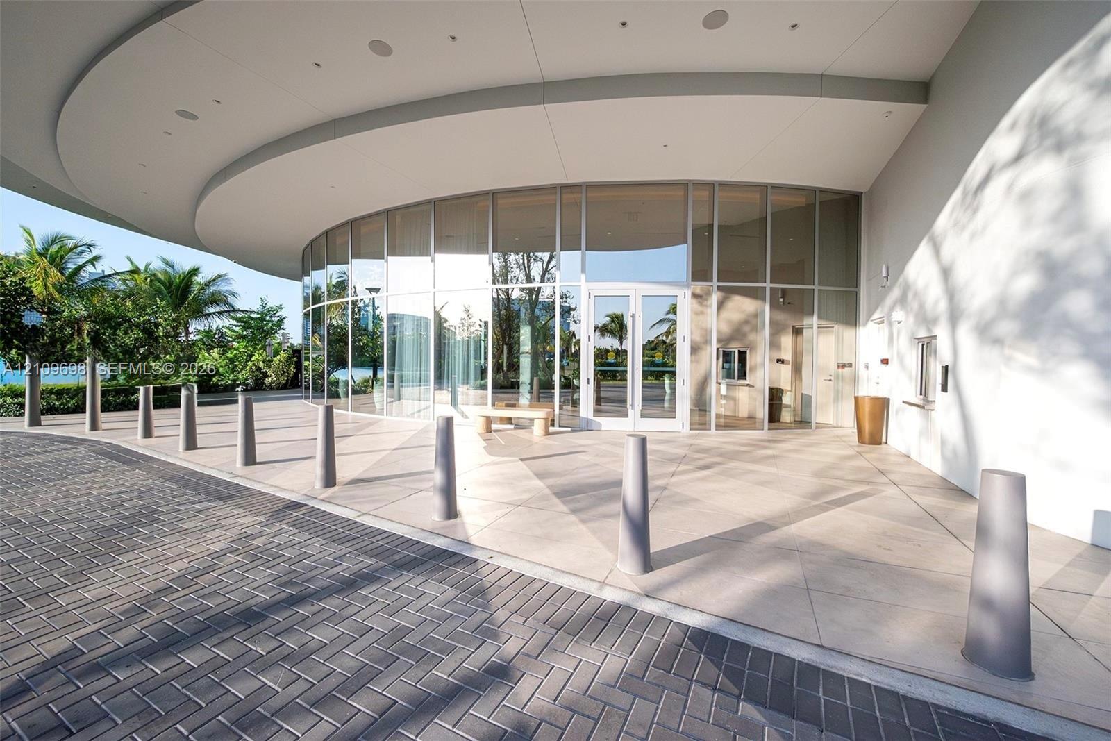 Curved glass faade featuring floor-to-ceiling windows and a deep structural overhang, complemented by a herringbone-patterned paver driveway and a wide concrete pedestrian plaza