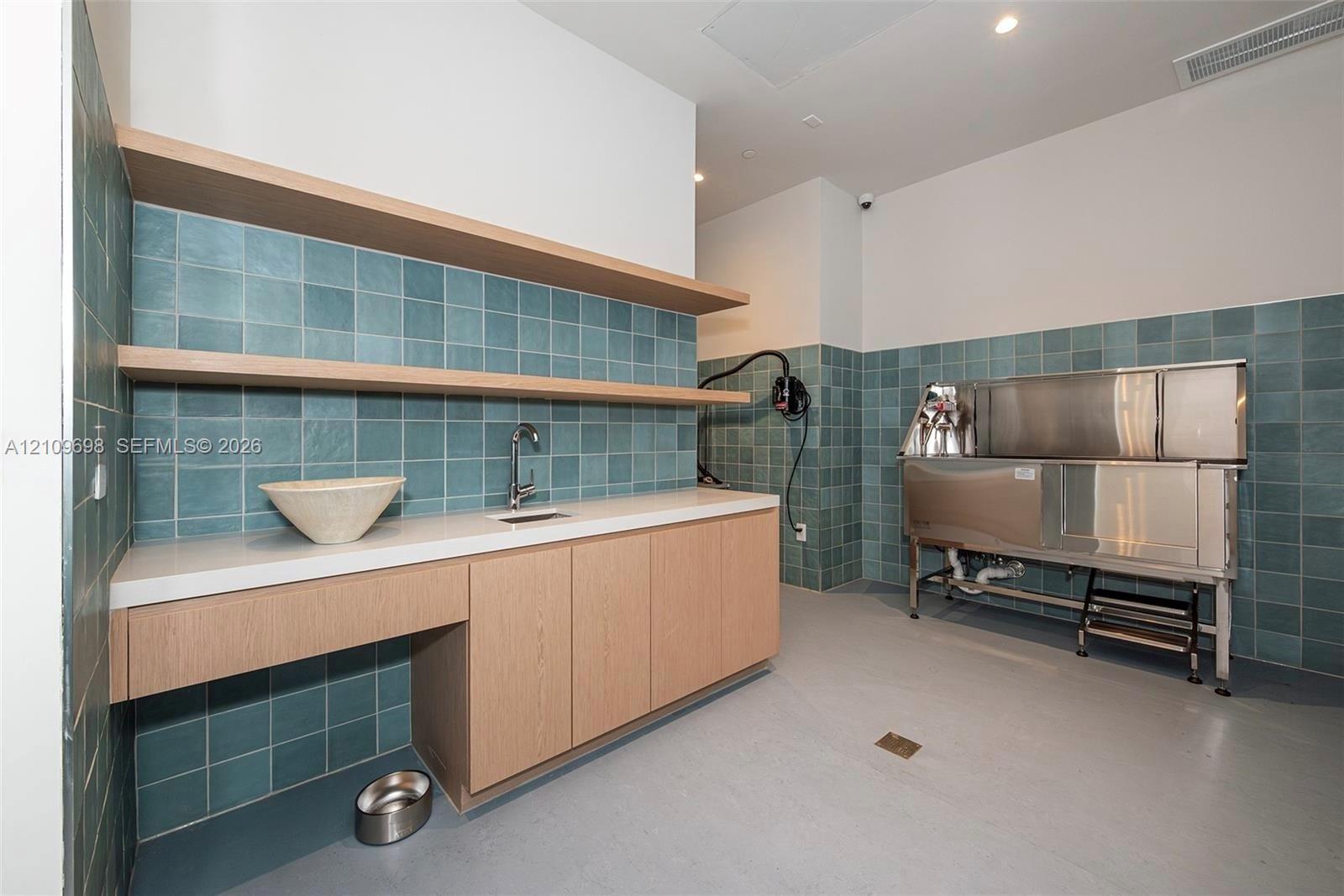 Utility space featuring custom wood cabinetry, quartz countertops, blue square tile wall backsplash, open floating shelving, and a stainless steel washing station