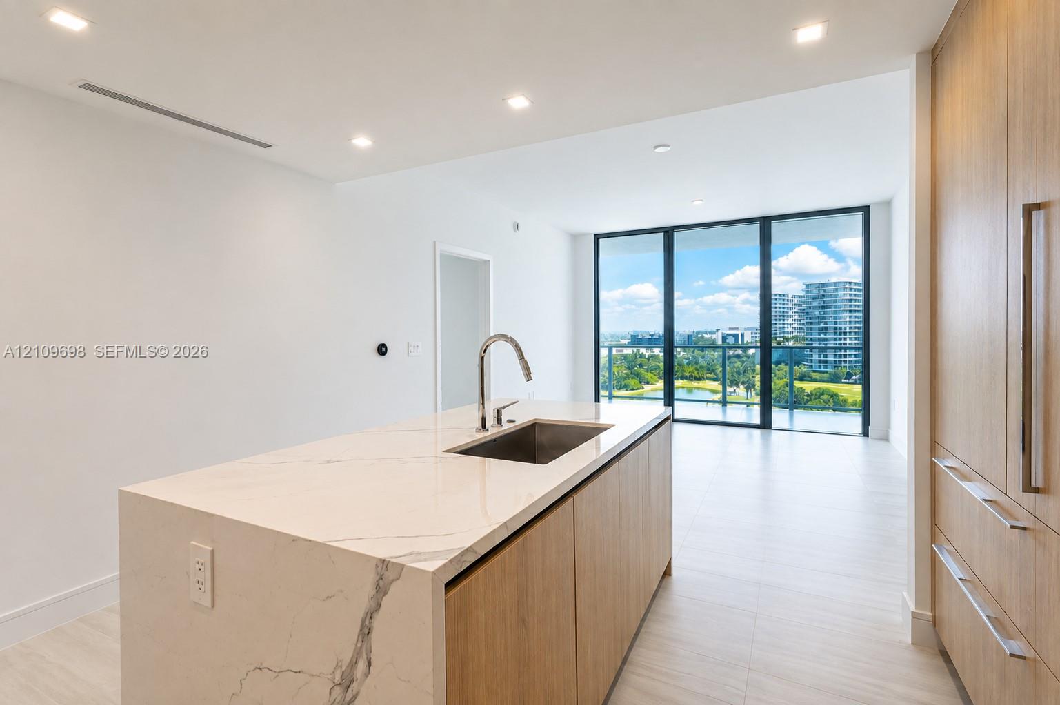 Kitchen island with stone-finish waterfall countertop, undermount stainless steel sink, wood-finish cabinetry, recessed lighting, and floor-to-ceiling glass paneling