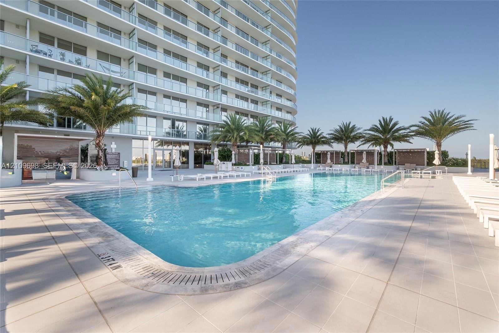 Large swimming pool with tiled surround, palm tree landscaping, and built-in outdoor kitchen area