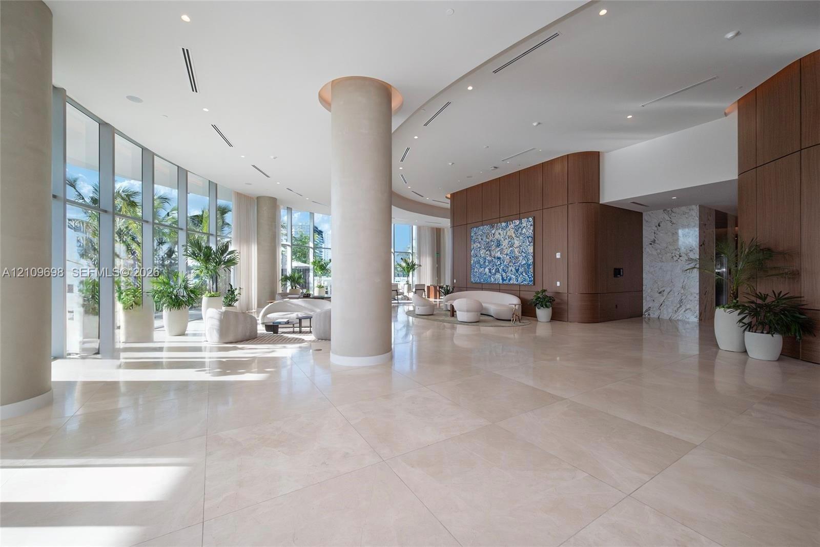 Expansive interior space featuring floor-to-ceiling windows, polished stone flooring, curved architectural pillars, wood-panelled accent walls, and recessed ceiling lighting