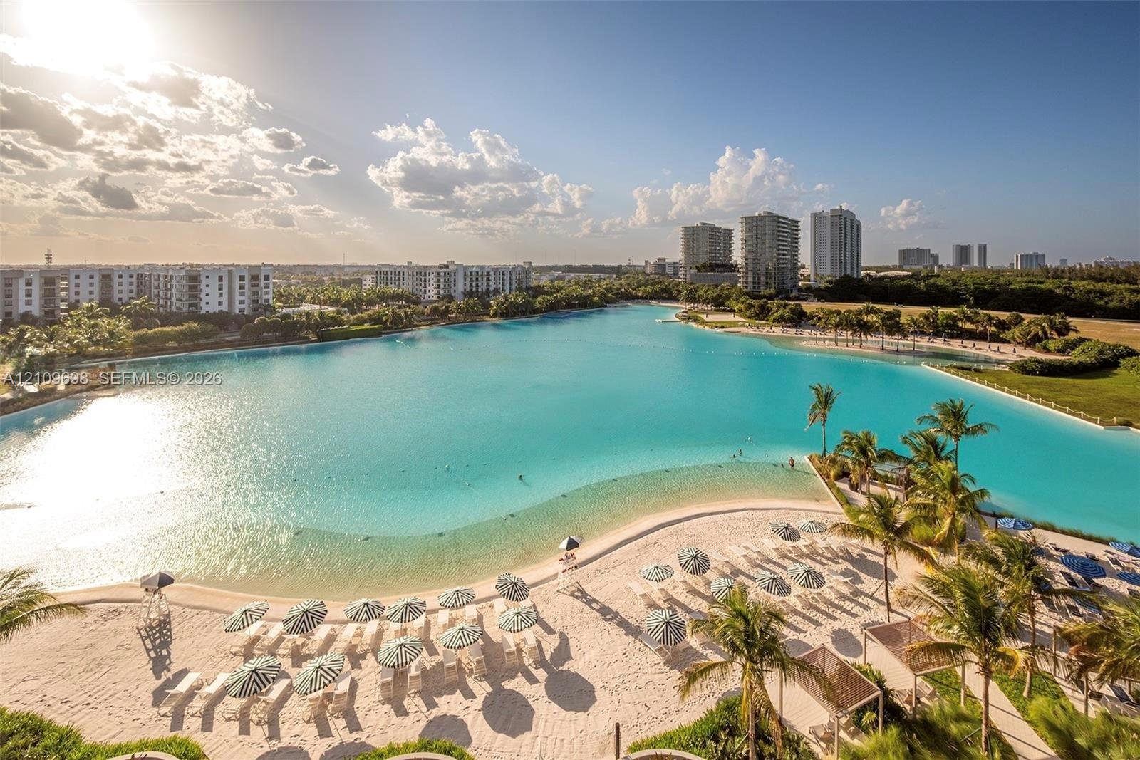 Extensive turquoise lagoon featuring a white sand beach, surrounding palm trees, and structural shaded areas