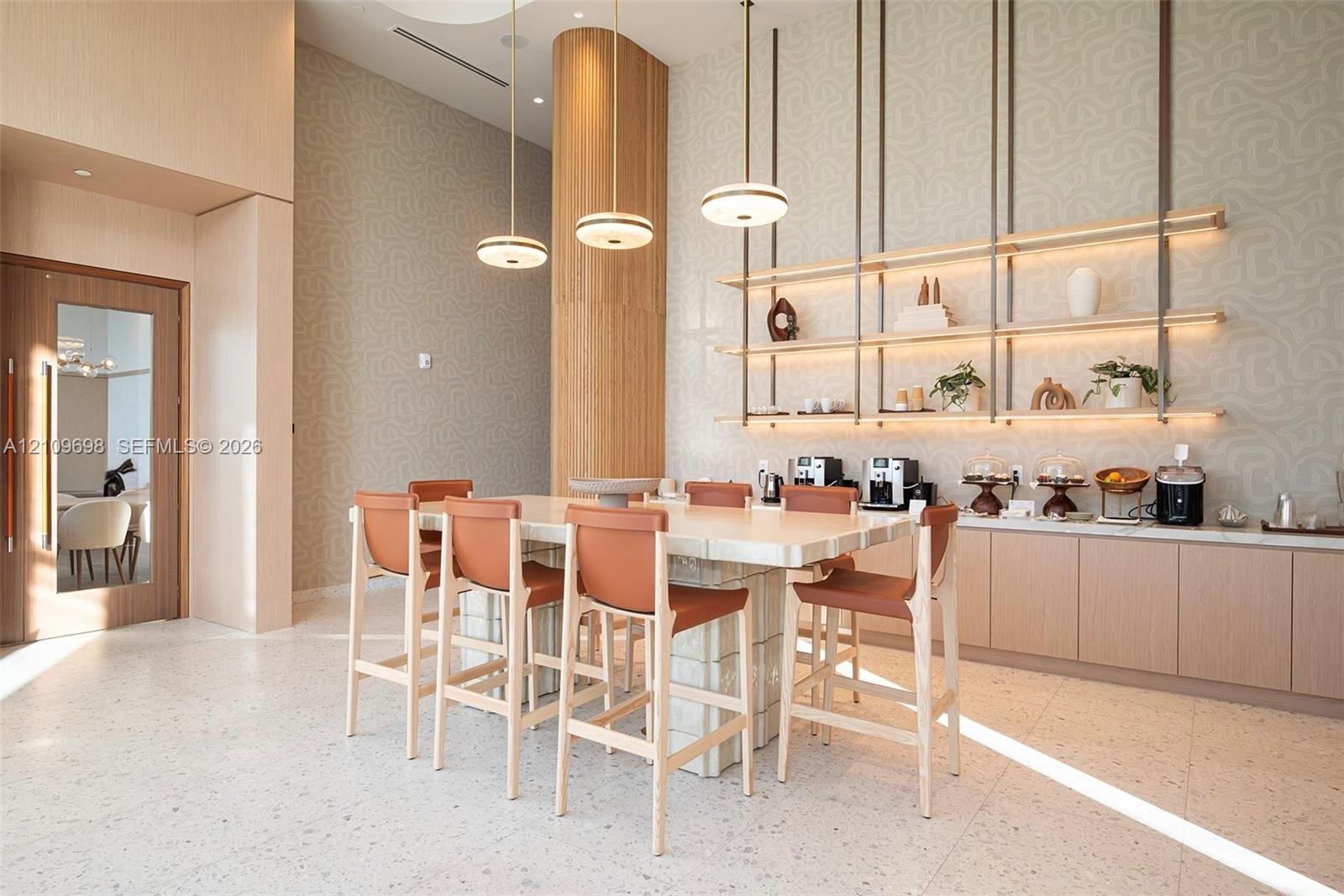 High-ceiling common area featuring terrazzo-style flooring, a central island with a tiled base, integrated wall-mounted shelving with recessed lighting, and modern multi-tiered pendant fixtures