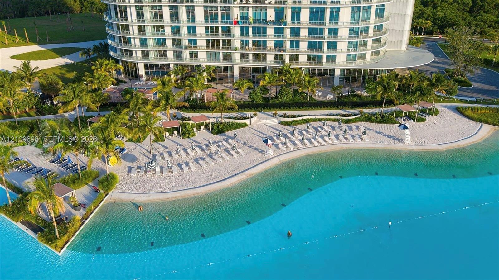 Aerial perspective of high-rise structure featuring a crystalline swimming lagoon, expansive sand beach, paved pathways, mature palm landscaping, and dedicated shaded cabanas