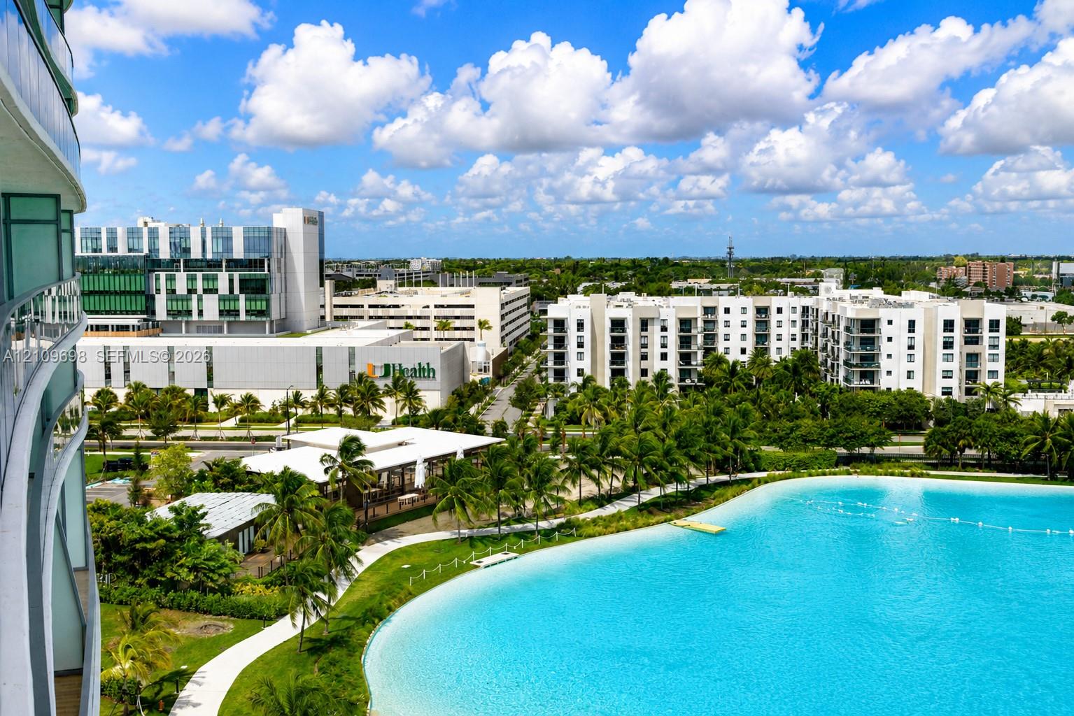 Elevated balcony featuring a glass railing, overlooking a large turquoise lagoon, landscaped grounds, palm trees, and paved walking paths