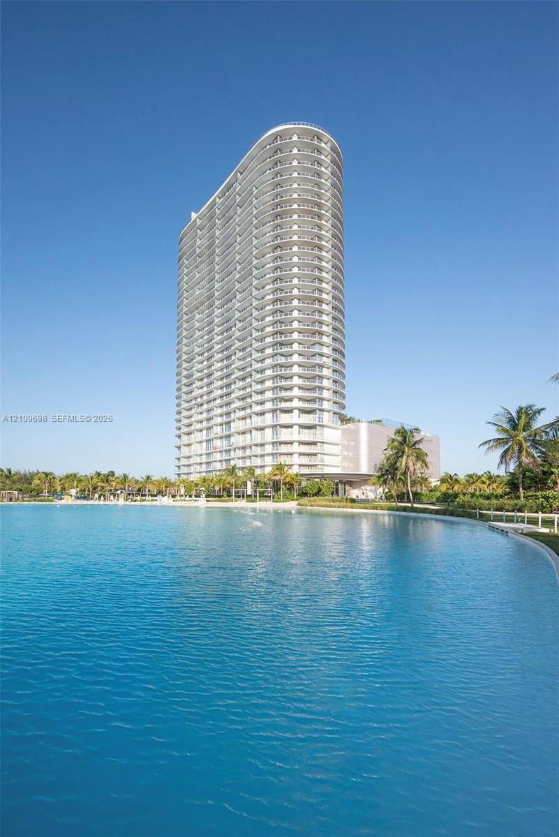 High-rise residential tower featuring curved architectural design, expansive wrap-around balconies, and floor-to-ceiling glass, situated on a waterfront lot with an artificial lagoon and landscaped palms