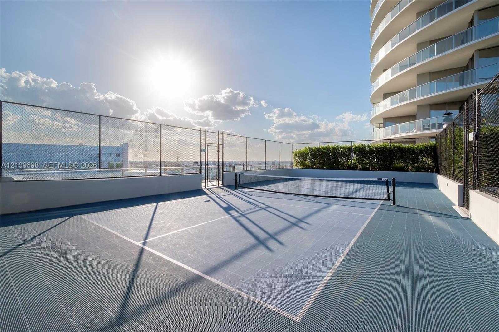Rooftop tennis court featuring a modular tile surface, perimeter chain-link fencing, and a built-in privacy hedge