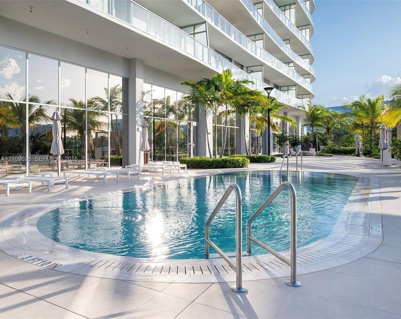 Swimming pool with metal handrails, expansive tile pool deck, floor-to-ceiling glass wall facade, and tropical landscaping