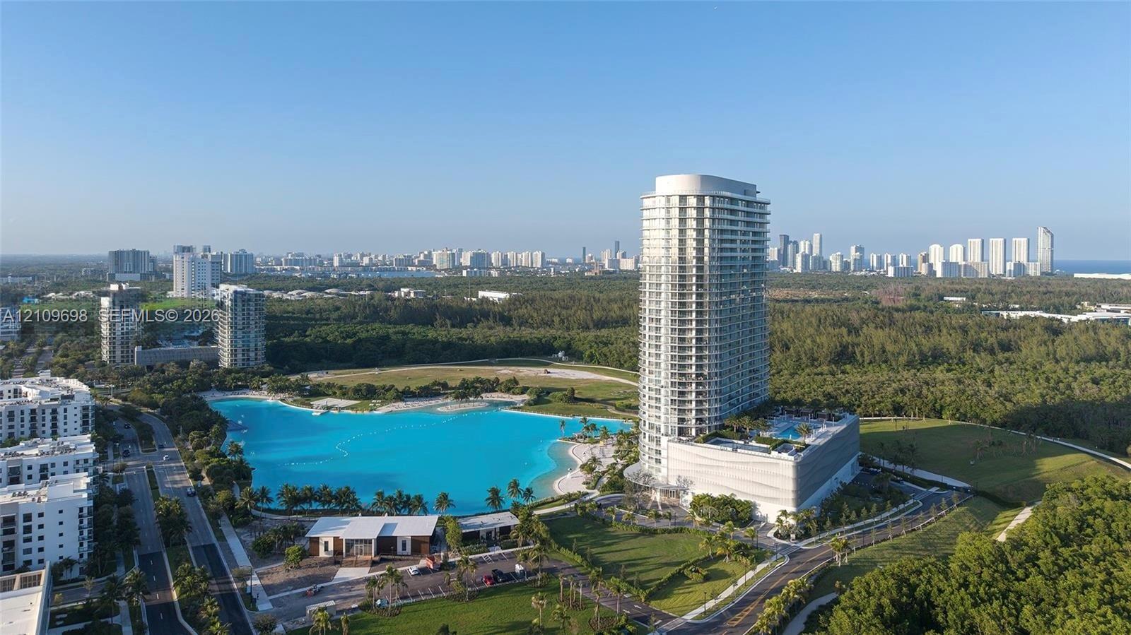 High-rise tower featuring extensive glass balcony railings, a large-scale lagoon-style pool, surrounding landscaped grounds, and a concrete parking structure