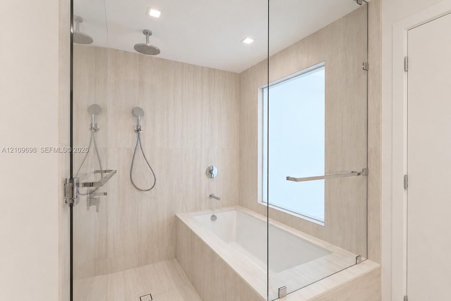Enclosed wet room featuring a deep soaking tub, frameless glass partition, dual wall-mounted rainfall showerheads, and neutral-toned stone-finish tiling