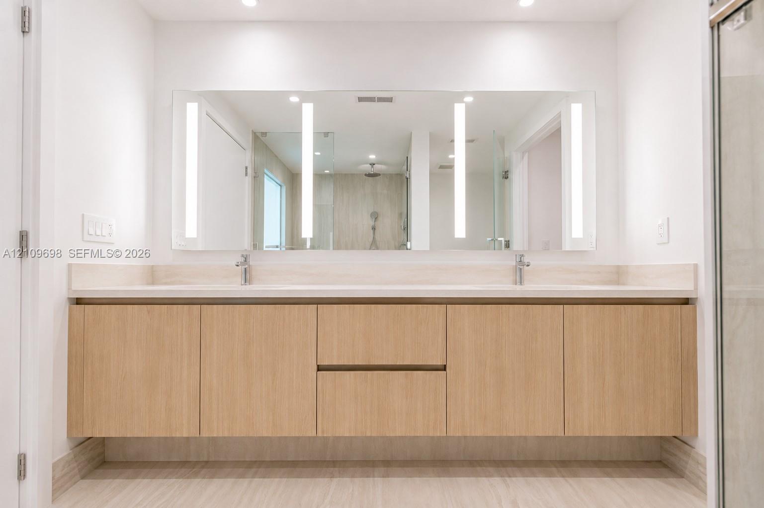 Floating dual-sink vanity featuring light wood-finish cabinetry, solid surface countertop, integrated LED-lit vanity mirror, and polished chrome fixtures