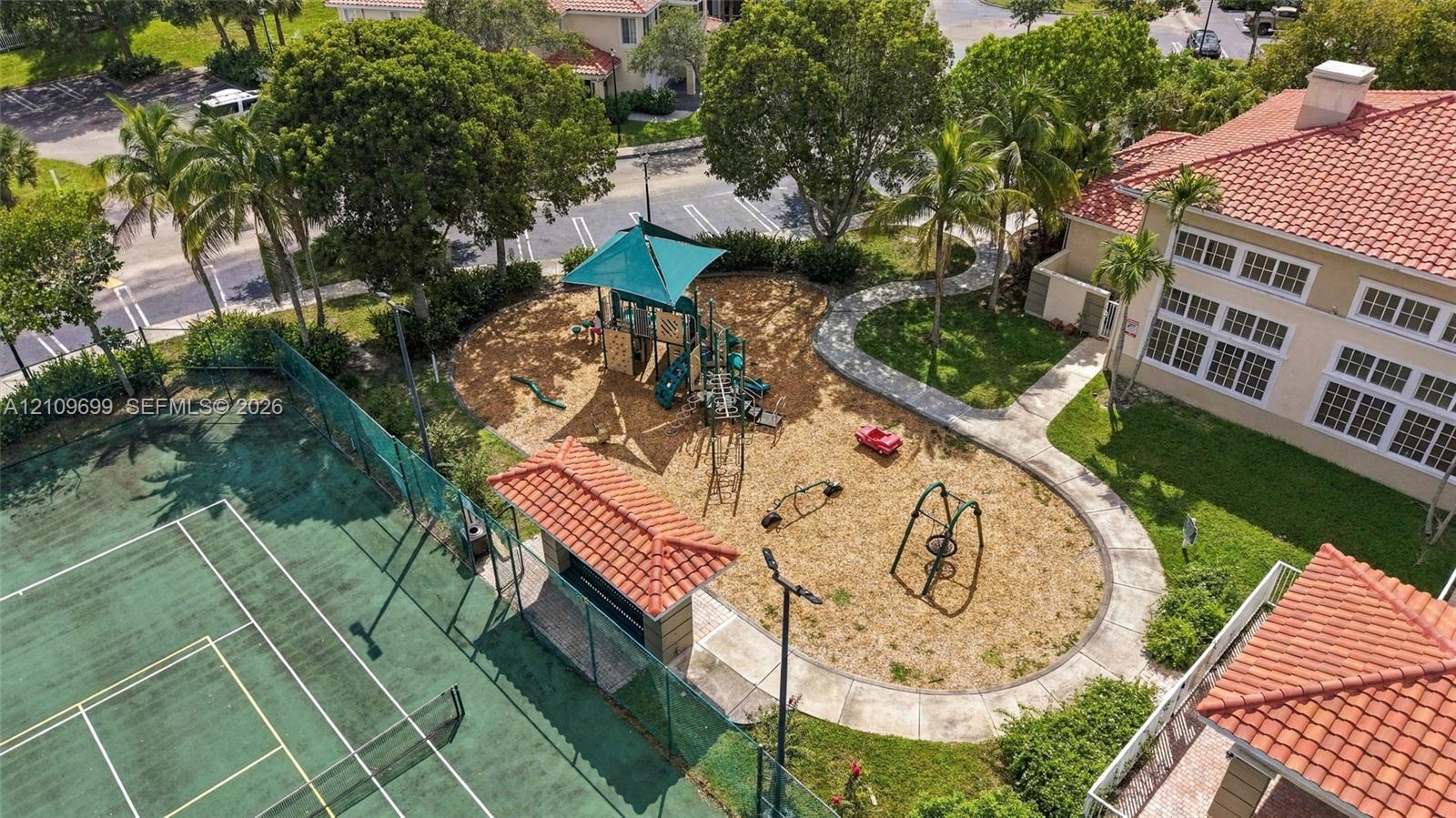 Recreational area featuring a fenced tennis court, shaded playground equipment, and a covered pavilion with a tiled roof