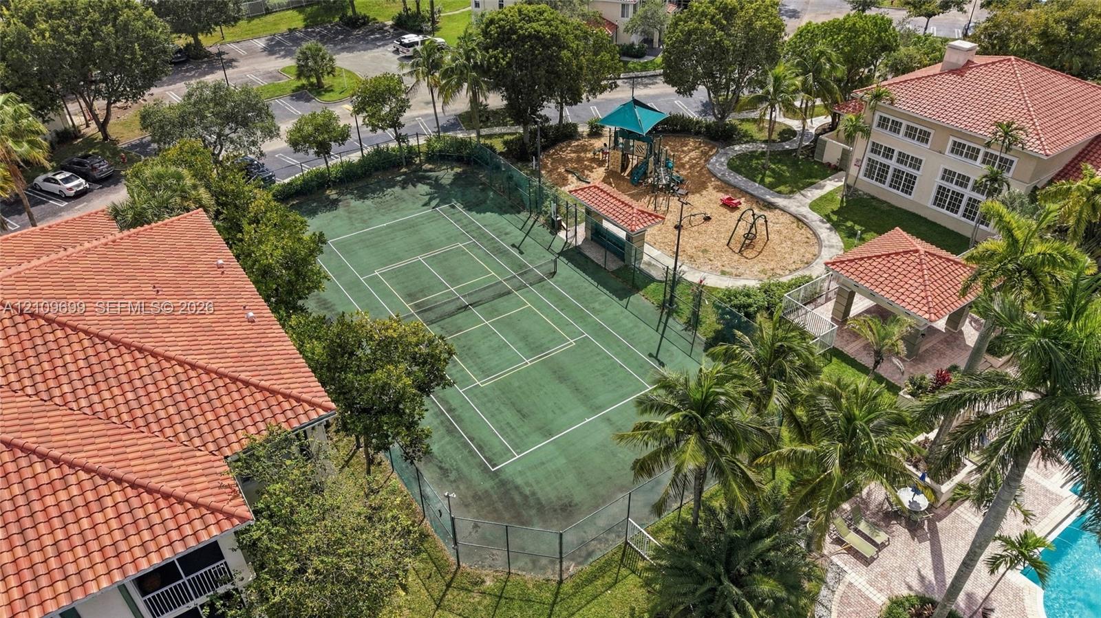 Tennis court with perimeter fencing, outdoor play area with structures, covered pavilion, and surrounding palm trees