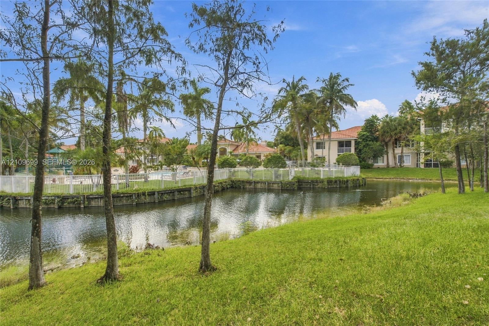 Waterfront property featuring a manicured lawn, mature palm trees, and a perimeter security fence