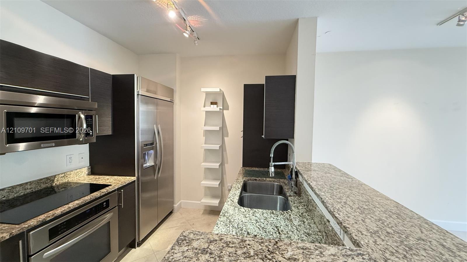 Kitchen featuring dark cabinetry, stone-finish countertops, stainless steel appliances, a double-basin undermount sink, and built-in vertical shelving Kitchen featuring dark cabinetry, stone-finish countertops, stainless steel appliances, a double-basin undermount sink, and built-in vertical shelving