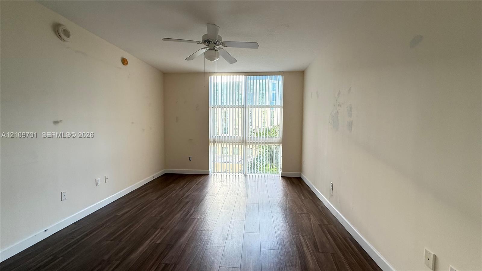 Room featuring wood-finish flooring, a ceiling fan with integrated lighting, and a floor-to-ceiling window assembly with vertical blinds Room featuring wood-finish flooring, a ceiling fan with integrated lighting, and a floor-to-ceiling window assembly with vertical blinds