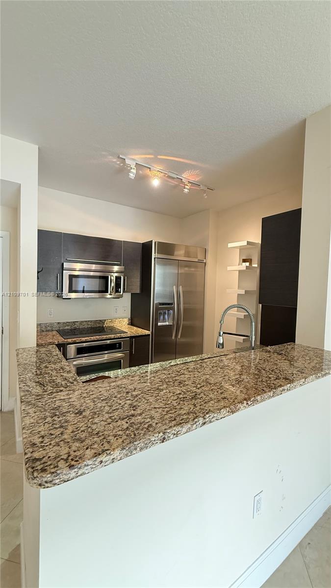 Kitchen featuring stone countertops, stainless steel appliances, dark cabinetry, track lighting, and built-in shelving Kitchen featuring stone countertops, stainless steel appliances, dark cabinetry, track lighting, and built-in shelving