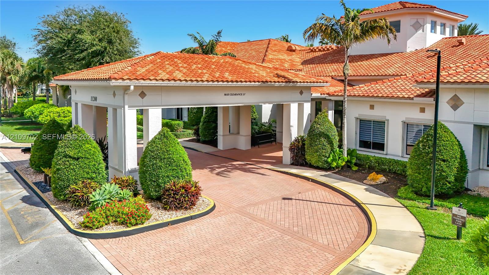 Mediterranean-style exterior featuring a covered porte-cochere, clay tile roofing, structured landscaping, and a brick-paver circular driveway