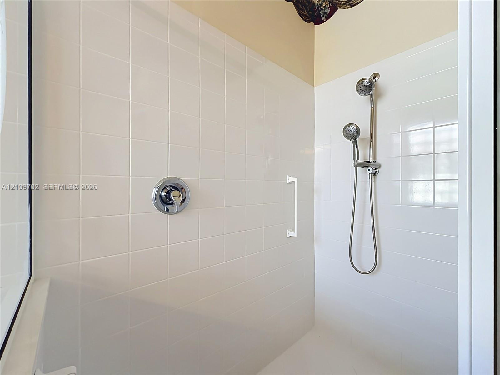 Shower enclosure featuring white square wall tiling, a chrome single-handle pressure balance valve, a dual-head shower system with a handheld attachment, and a white grab bar