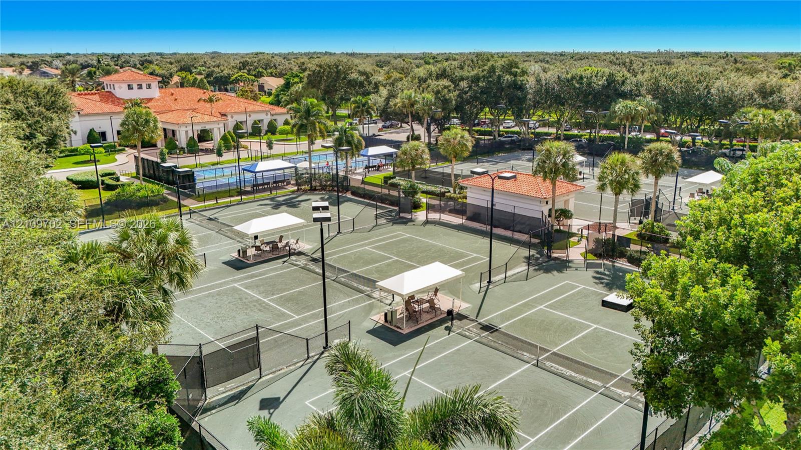 Aerial view of multiple fenced tennis courts featuring hard-court surfaces, elevated lighting, and shaded seating structures