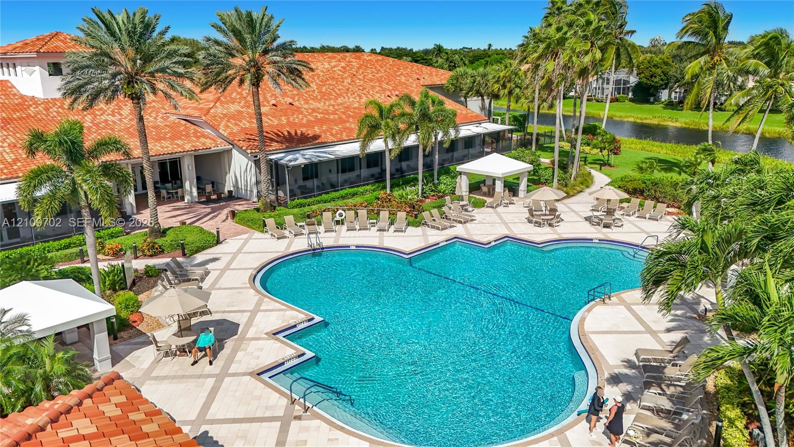 In-ground pool with expansive stone-paved deck, tile-roofed clubhouse, and mature palm landscaping