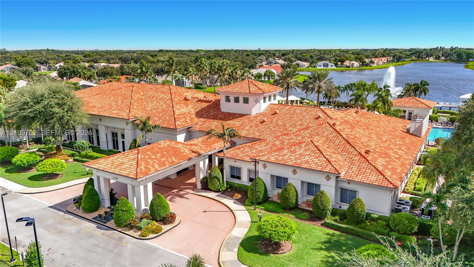 Expansive clubhouse featuring a terracotta tile roof, covered porte-cochre entry, surrounding manicured landscaping, and exterior water feature