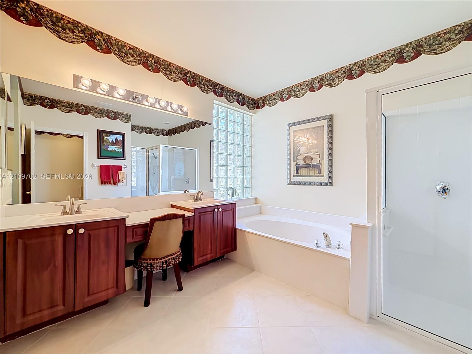 Bathroom featuring dual-sink vanity with wood-finish cabinetry, glass block window, deep soaking tub, and enclosed glass shower