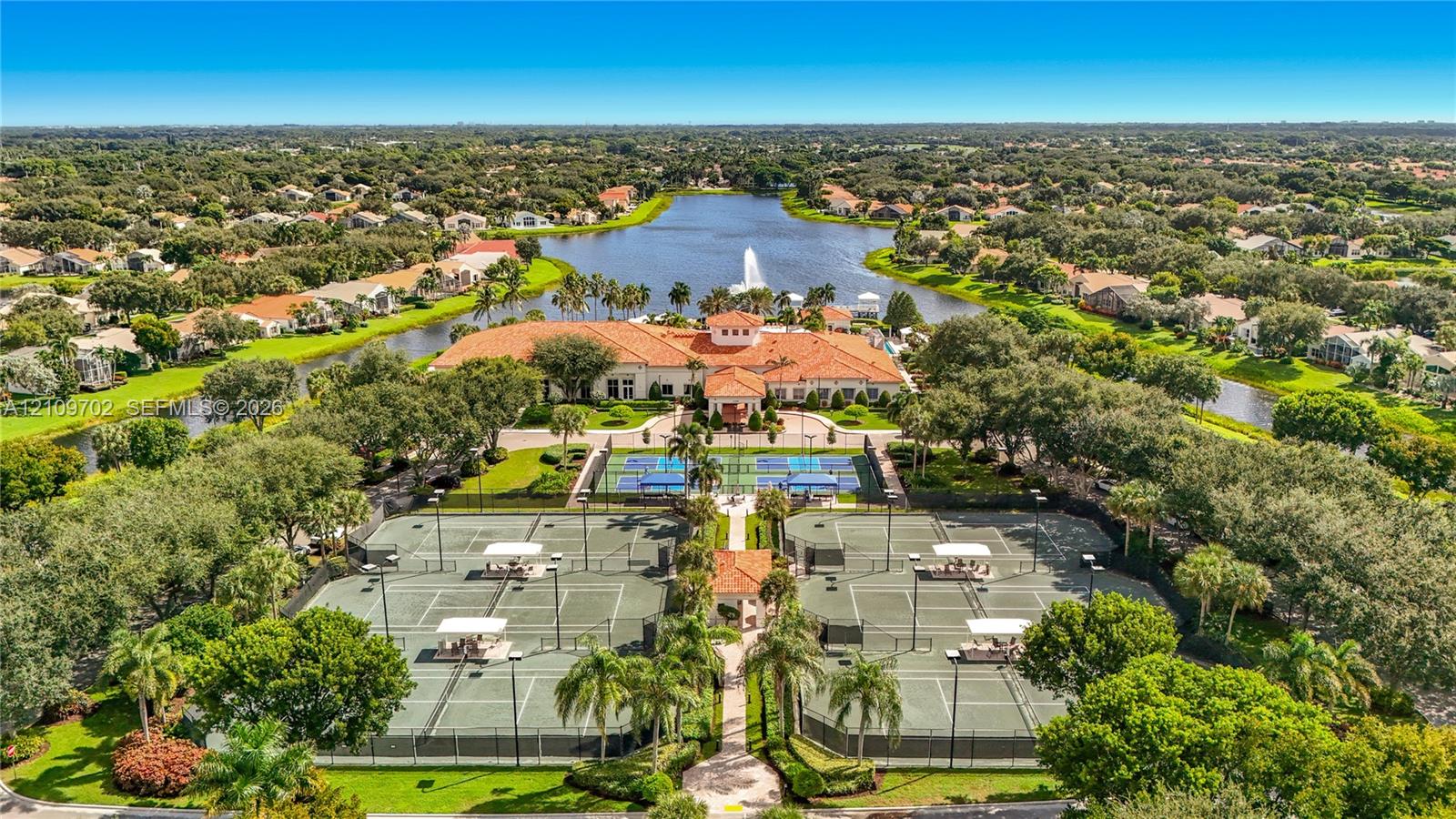Aerial perspective of a community facility featuring multiple tennis courts, a large central building with a tile roof, and a water feature with a fountain