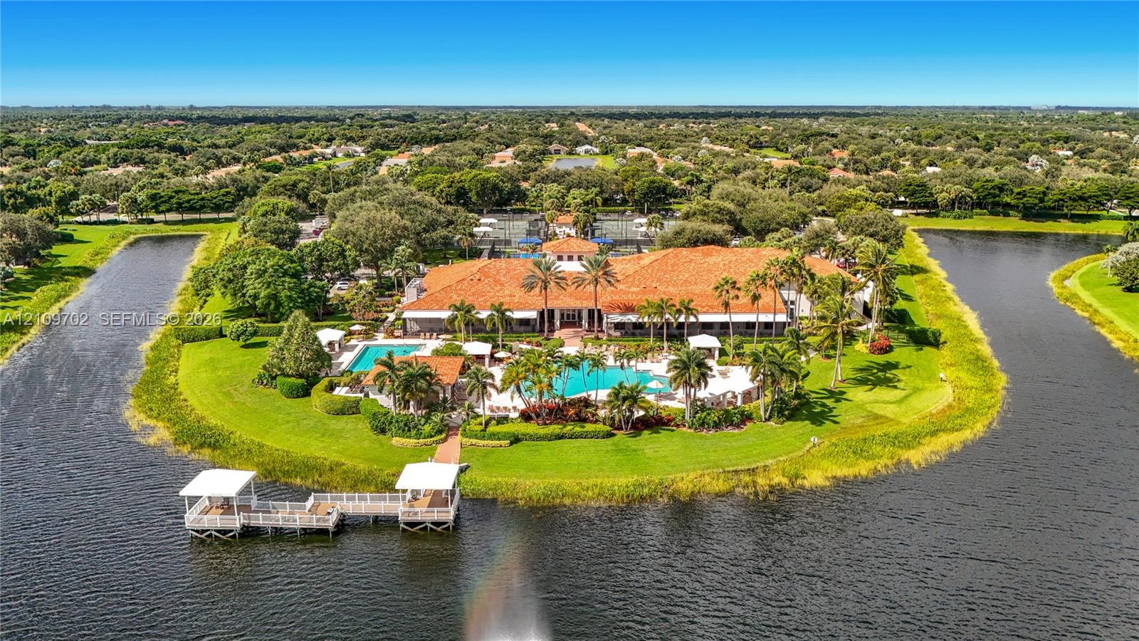 Waterfront Clubhouse featuring a terra cotta tile roof, multiple outdoor swimming pools, a private wooden dock, and extensive landscaped grounds